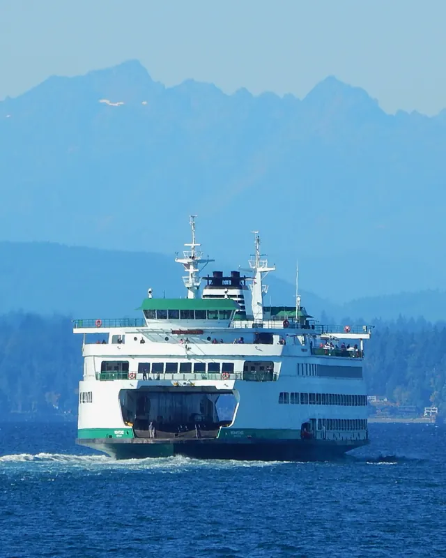 A white and green Washington State Ferries boat crosses Puget Sound with the snowy Olympic Mountains behind.